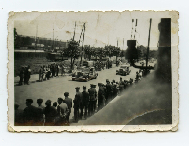 Badauds et quelques militaires le long de la route regardant le passage de véhicules. Foule dans une tribune.