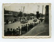 Badauds et quelques militaires le long de la route regardant le passage de véhicules. Foule dans une tribune.