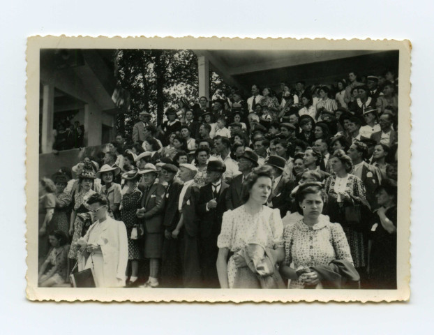 Badauds et quelques militaires le long de la route regardant le passage de véhicules. Foule dans une tribune.