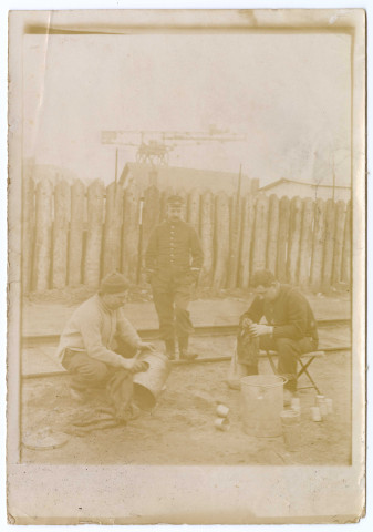 Trois hommes en extérieur dont deux assis en train de nettoyer des gamelles